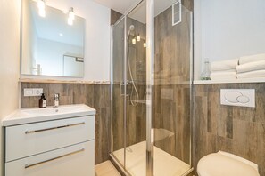 Um banheiro moderno com chuveiro de vidro, pia branca com dosador de sabão e vaso sanitário.