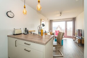 Eine moderne Küche mit Holzarbeitsplatte, einem Toaster und einer Kaffeemaschine. Ein Essbereich mit einem rosa Stuhl und einem Tisch. Der Raum hat einen Balkon mit Blick.