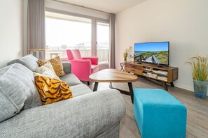 A modern living room with a grey sofa, a pink chair, a wooden coffee table, and a TV on a wooden stand.