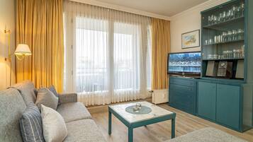 Un salon comprenant un canapé, une table basse et un téléviseur. On y trouve également un meuble contenant de la verrerie et un tableau accroché au mur.