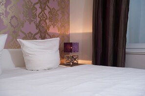 A neatly made bed with white linens, a single white pillow, and a small table with a purple lamp beside it.