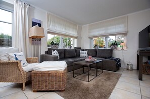 Ein modernes Wohnzimmer mit grauem Sofa, Korbstühlen, einem Couchtisch und großen Fenstern mit Vorhängen.