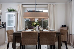 Ein Esszimmer mit einem für vier Personen gedeckten Tisch, umgeben von Korbstühlen, einer Vitrine mit Glastüren und einem Fenster mit Vorhängen.