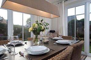 A dining table set with white plates, silverware, and wine glasses, surrounded by wicker chairs. A vase with flowers is placed on the table, and a radio is visible in the background.
