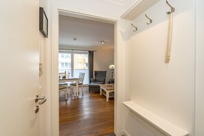 Un pasillo que conduce a una sala de estar con un sofá, una mesa y una lámpara. Hay una puerta con un perchero y un marco de cuadro en la pared.