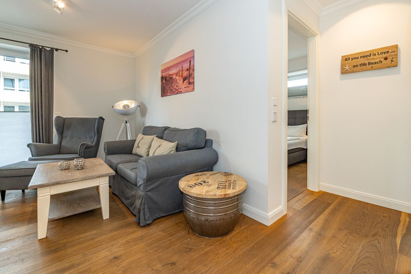 A modern living room with a grey sofa, a small table, and a lamp. There is a wooden barrel and a painting on the wall.