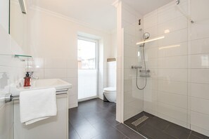 Un baño moderno con cabina de ducha de vidrio, un lavabo blanco con dispensador de jabón y un inodoro visible a través de una ventana.