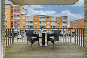 Un balcón con dos sillas de mimbre y una mesa, con vista a un edificio de varios pisos con baldosas anaranjadas y blancas.