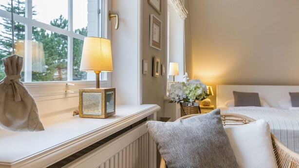 A hotel room with a bed, a window with a view of trees, a bedside table with a lamp, and a radiator. - Sönshörn Suite - 2-room vacation apartment on the Wadden Sea, in Munkmarsch. (Sylt)