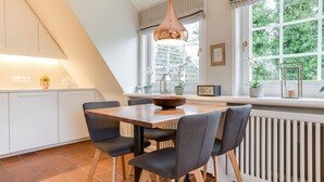 A modern kitchen with a dining area, featuring a large window with a view of greenery, a pendant light, and a radio. - Sönshörn Suite - 2-room vacation apartment on the Wadden Sea, in Munkmarsch. (Sylt)