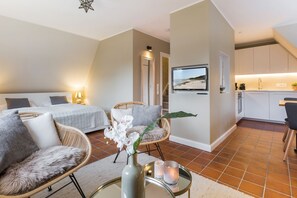 A modern hotel room with two single beds, a sitting area with wicker chairs, a small table with a vase and candle, and a kitchen area with a TV displaying a beach scene.