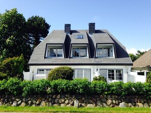 A two-story house with a stone wall in front, a large window on the ground floor, and a chimney on the roof.