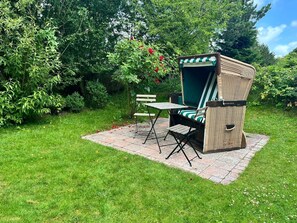 Un patio jardín con una silla de mimbre y una mesa, rodeado de abundante vegetación y un camino de piedra.