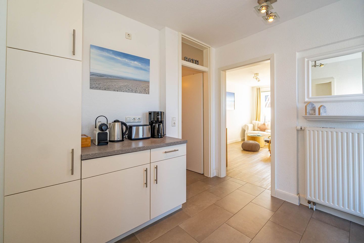 Eine moderne Küche mit weißen Schränken, einer Kaffeemaschine, einem Wasserkocher und einem Toaster auf der Arbeitsfläche. Darüber an der Wand hängt ein Gemälde eines Strandes.