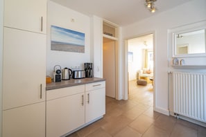 A modern kitchen with white cabinets, a coffee maker, a kettle, and a toaster on the countertop. There is a painting of a beach on the wall above the counter.