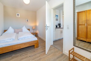 A bedroom with a wooden bed, a nightstand, a bedside table, a mirror, and a view into the kitchen.