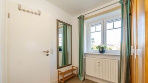 A room with a white door, a mirror, a window with green curtains, a radiator, and a small wooden bench. - Marienhüs - Cozy, central vacation apartment in Westerland for up to 2 (Sylt)