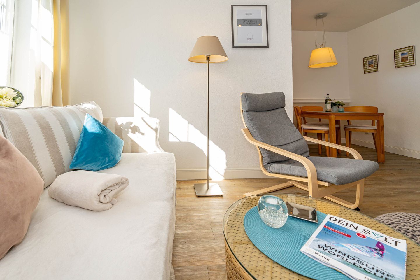 Ein Wohnzimmer mit einer Couch, einem grauen Sessel, einer Stehlampe, einem Esstisch mit Stühlen und einer Zeitschrift auf einem Beistelltisch.