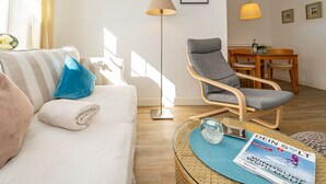 A living room with a sofa, a grey armchair, a floor lamp, a dining table with chairs, and a magazine on a side table. - Marienhüs - Cozy, central vacation apartment in Westerland for up to 2 (Sylt)