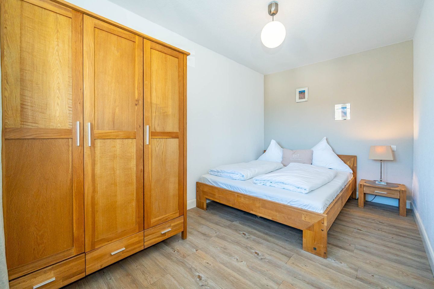 Ein Schlafzimmer mit Holzschrank, einem Bett mit weißer Bettwäsche, einem Nachttisch mit Lampe und einem kleinen Bild an der Wand.