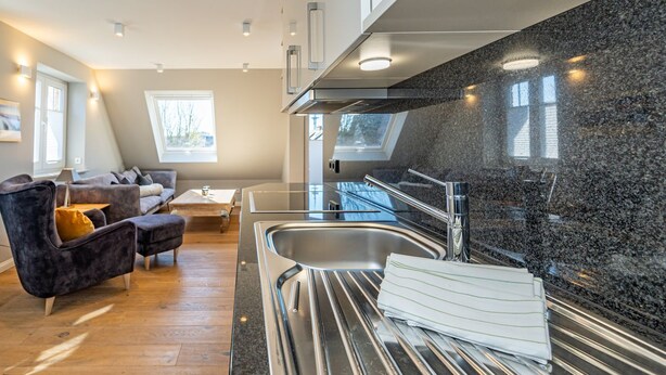 A modern kitchen with a granite countertop, stainless steel sink, and a built-in oven. There is a dining area with a dark sofa and a small table. - Sturmhaube - Apartment for max. 4 persons in Alt-Westerland with balcony. (Sylt)