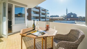 A balcony with a table, wicker chair, and a bottle of wine. - Strandnest - Stylische, Neue Wohnung in Strandnähe (Sylt)