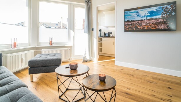 A modern living room with a grey sofa, two round tables with stools, a large window with curtains, and a painting on the wall. - Strandnest - Stylische, Neue Wohnung in Strandnähe (Sylt)