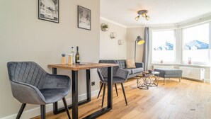 A modern living room with a wooden dining table, two grey upholstered chairs, a grey sofa, and a small grey ottoman. There are framed pictures on the wall, a floor lamp, and a window with a view of buildings. - Strandnest - Stylische, Neue Wohnung in Strandnähe (Sylt)