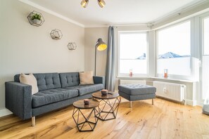 A modern living room with a grey sofa, two round tables with geometric legs, a floor lamp, and a large window with a view of buildings.