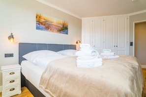 Un lit bien fait, recouvert d’une couverture beige, avec des oreillers blancs et des serviettes pliées, le tout posé sur un plancher en bois.