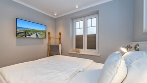 Une chambre avec deux lits, une fenêtre avec des volets, un téléviseur fixé au mur et une petite étagère ornée d’objets décoratifs.