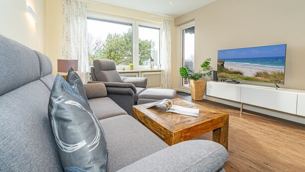 Ein modernes Wohnzimmer mit einem grauen Sofa, einem Holztisch und einem Fernseher, der eine Strandlandschaft zeigt.