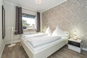 A bedroom with a bed, a nightstand, a window with curtains, a radiator, and a patterned wallpaper.