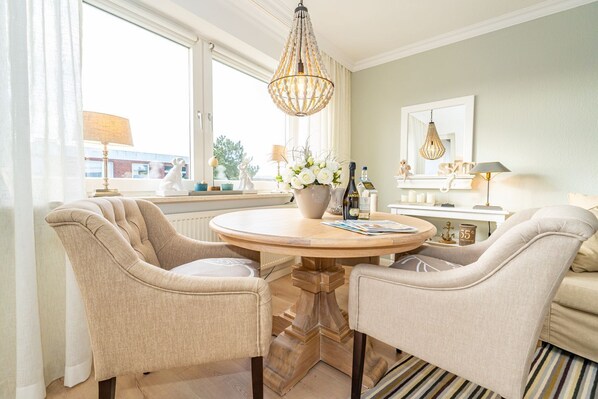 Un coin repas comprenant une table ronde, deux chaises rembourrées, un lustre et une fenêtre avec des rideaux.