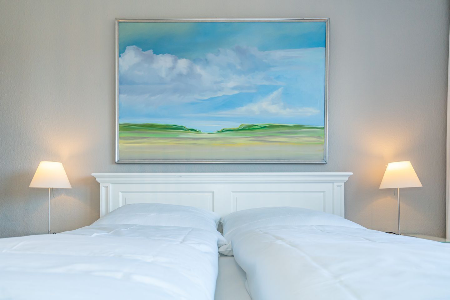 A hotel room with two beds, a white headboard, and a painting of a landscape above the beds.