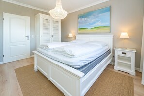Ein ordentlich bezogenes Bett mit weißen Leinen, ein weißer Nachttisch mit einer Lampe und ein Landschaftsbild an der Wand.