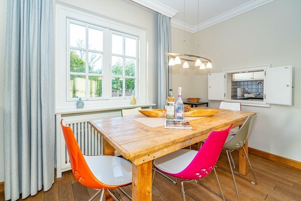 A dining area with a wooden table, four chairs, a window with curtains, a hanging light fixture, and a kitchen visible through an open door.