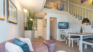 A modern living room with a pink sofa, a dining table with chairs, a staircase, and a TV in the background.