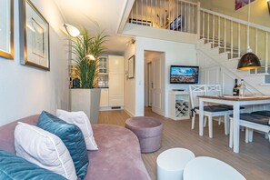A modern living room with a pink sofa, a dining table with chairs, a staircase, and a TV in the background.