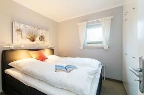 Ein ordentlich gemachtes Bett mit weißer Bettwäsche und einem Buch darauf, ein Nachttisch, eine Wandlampe, ein Fenster mit Vorhängen und eine Tür mit Griff.