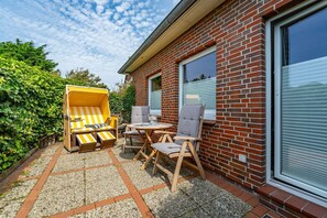 Un patio con mobiliario exterior, que incluye un sofá a rayas amarillas y dos sillas de madera, frente a un muro de ladrillo con ventanas grandes.