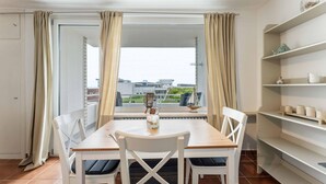 A dining area with a table and chairs, a view of buildings through a window, and a shelf with items. - Seestern - Maritime Wohnung mit Meerblick und Balkon (Sylt)