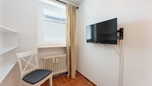 A small room with a white radiator, a window with blinds, a chair, and a mounted television. - Seestern - Maritime Wohnung mit Meerblick und Balkon (Sylt)