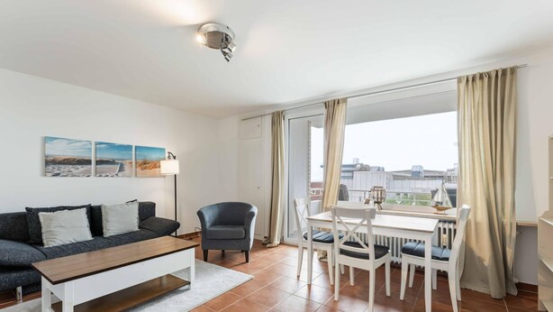 Een moderne woonkamer met een bank, salontafel, eettafel en stoelen. Er is een groot raam met gordijnen en kunst aan de muur.