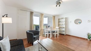 A modern living room with a sofa, a dining table with chairs, a bookshelf, and a large window with curtains. - Seestern - Maritime Wohnung mit Meerblick und Balkon (Sylt)