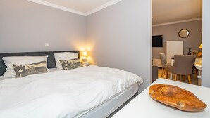 Una cama bien hecha con ropa de cama blanca y almohadas decorativas, una mesita de noche con una lámpara y un objeto de madera sobre una mesa.