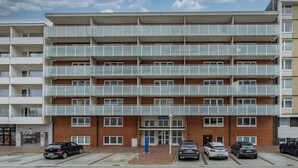 Un edificio residencial de varios pisos con fachada de ladrillo, múltiples balcones y un estacionamiento con varios coches.