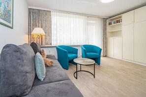 A modern living room with a grey sofa, two blue armchairs, a small round table, and a floor lamp. There is a bookshelf with books and a radiator under the window.