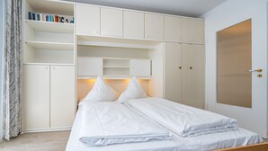 A bedroom with a bed, white bedding, a wooden headboard, a wardrobe with multiple compartments, and a door with a window. - Neptun - Wohnen am Meer, in Dieser Zentralen Wohnung (Sylt)
