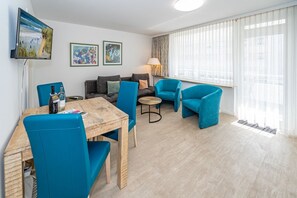 Ein modernes Wohnzimmer mit einem Holzesstisch, blauen Stühlen, einer Couch und einem an der Wand befestigten Fernseher. Über der Couch hängen zwei gerahmte Kunstwerke, und auf einem Beistelltisch steht eine Lampe.
